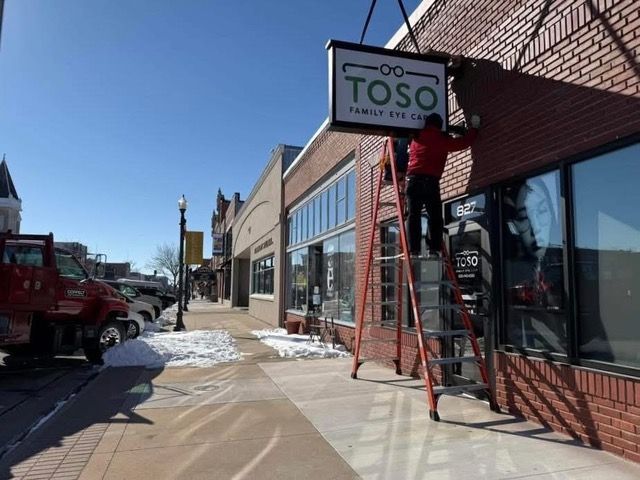 Man on ladder installing a business sign 'TOSO Family Eye Care' on a brick building in a snowy town.