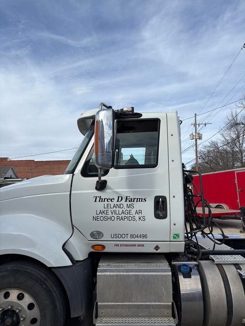 White semi-truck with Three D Farms logo, addresses in Leland, MS, Lake Village, AR, and Neosho Rapids, KS. Against a cloudy sky.