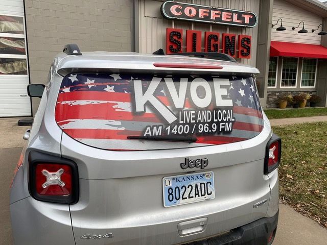 Silver Jeep with KVOE radio station logo and American flag design on the rear window, parked in front of a sign shop.