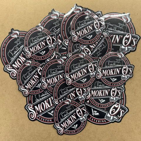 Pile of Smokin' O's Custom Catering stickers with a black and red design on a brown surface.