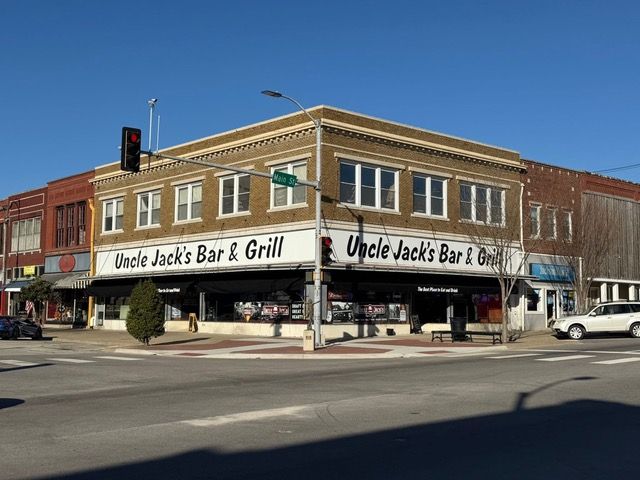 Uncle Jack's Bar & Grill is built on a corner, with brown brick and a black awning, a car, and a traffic light.
