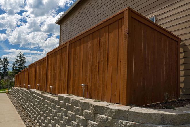 Brown wooden fence next to a retaining wall and sidewalk, under a blue sky with clouds.