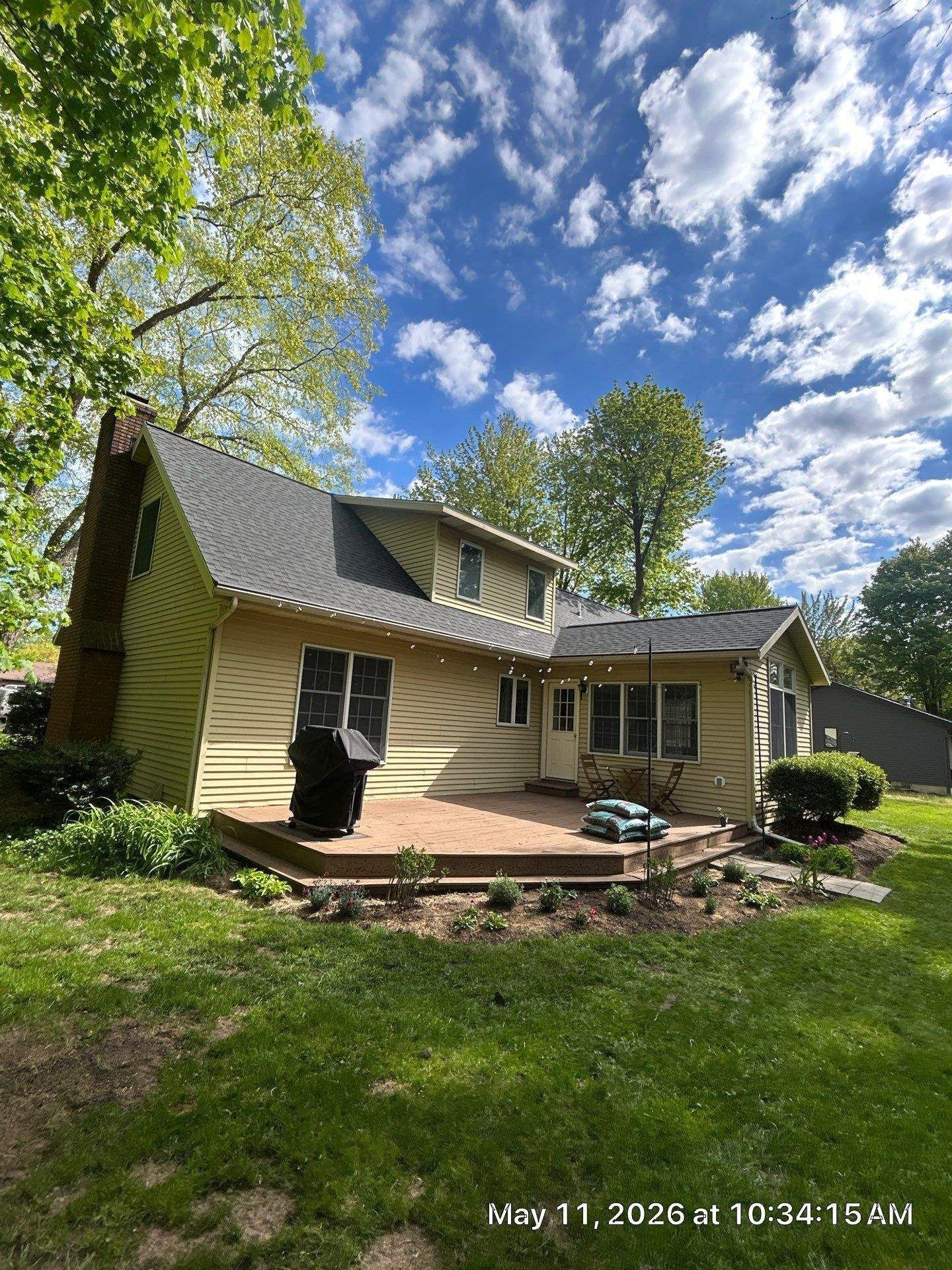 Suburban house with beige siding, dark roof, brick patio, and green lawn under a bright cloudy sky