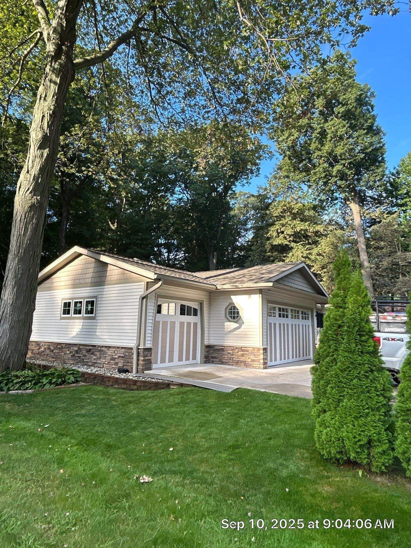 Garage with light siding and two garage doors, set against a backdrop of trees and lawn.