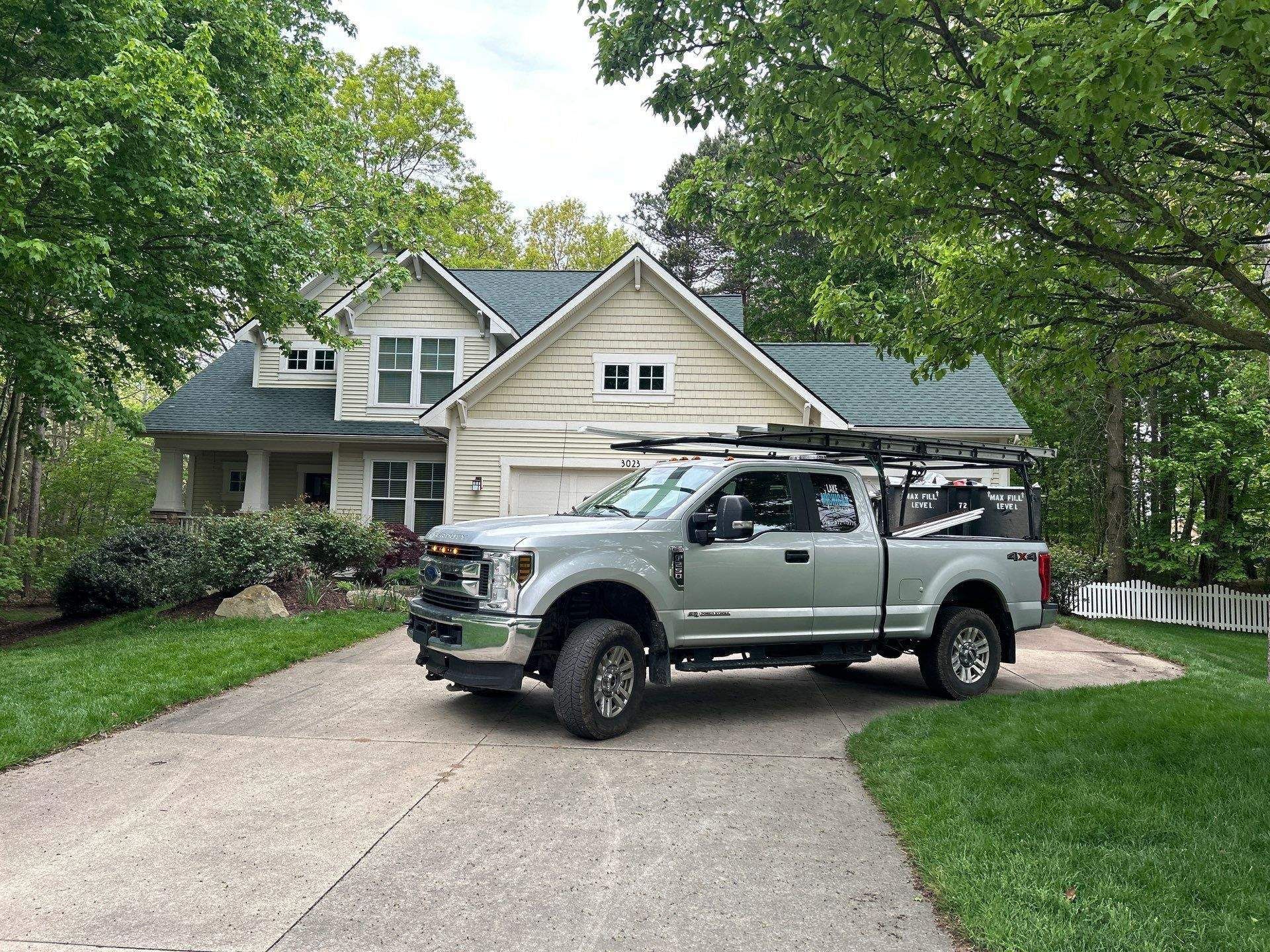 Silver pickup truck parked in front of a light-colored house on a driveway, trees surrounding.
