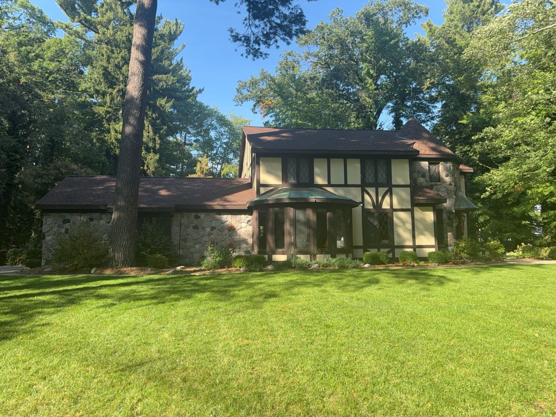 Tudor-style house with stone and timber facade, brown roof, and lush green lawn. Set in a wooded area.