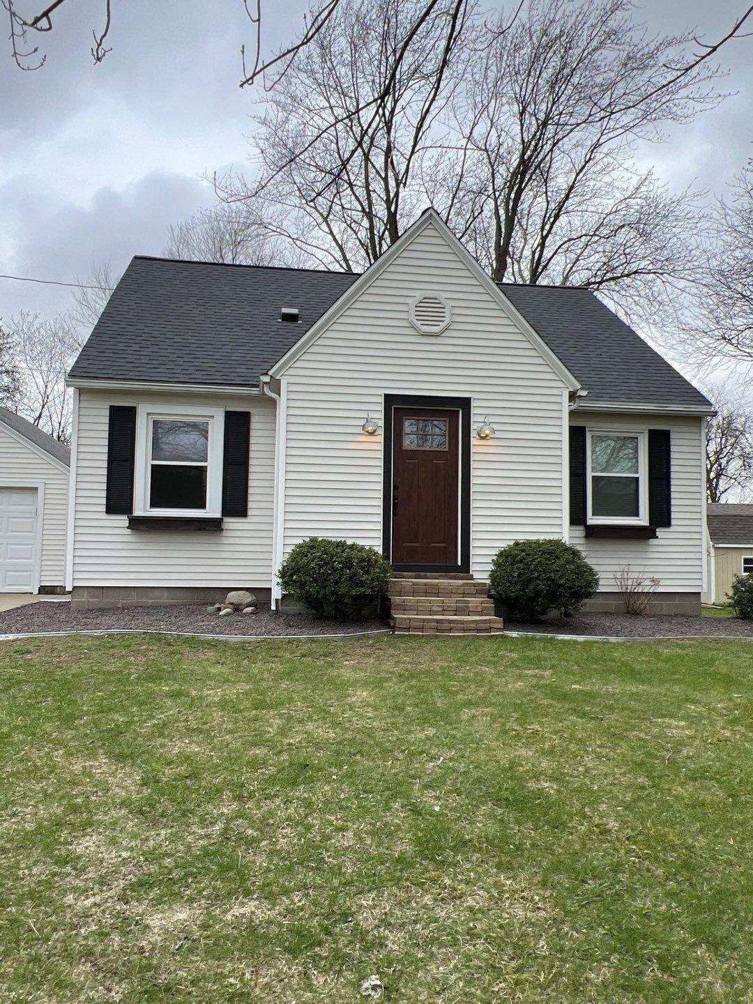 Small, white house with black shutters, dark roof, brown door, and green lawn.