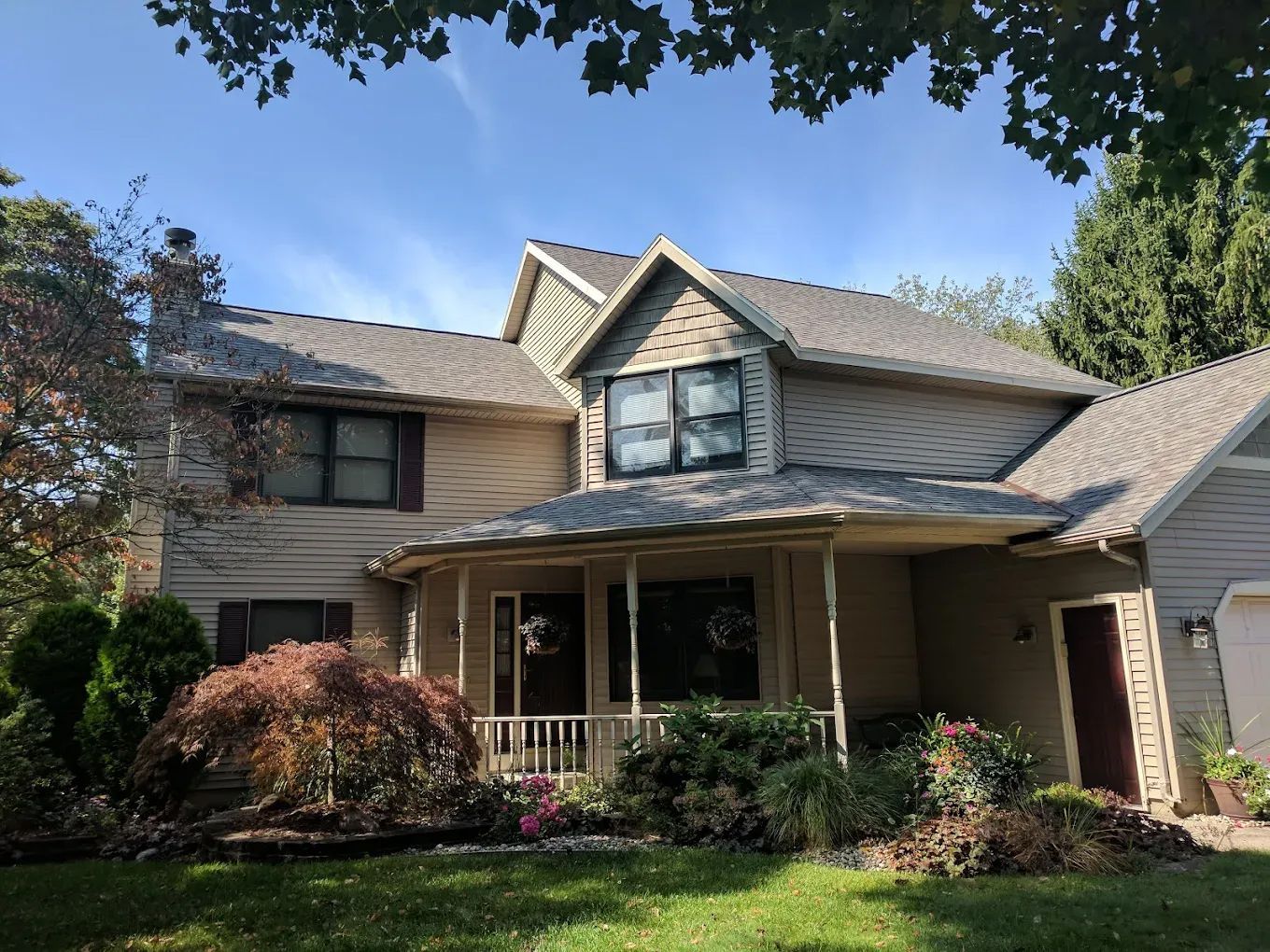 Two-story beige house with a porch, lush landscaping, and a blue sky.