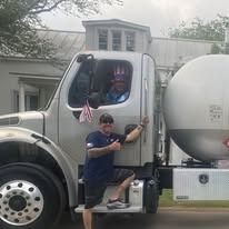 Man and truck driver giving thumbs up, truck with tank, flag, in front of a building.