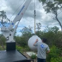 A crane lifting a large white tank; a person watches. Outdoors, cloudy sky.