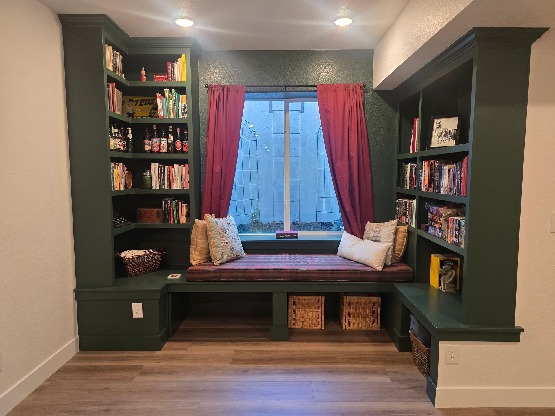 Cozy reading nook with built-in bookshelves painted dark green, a window with burgundy curtains, and a cushioned window seat.