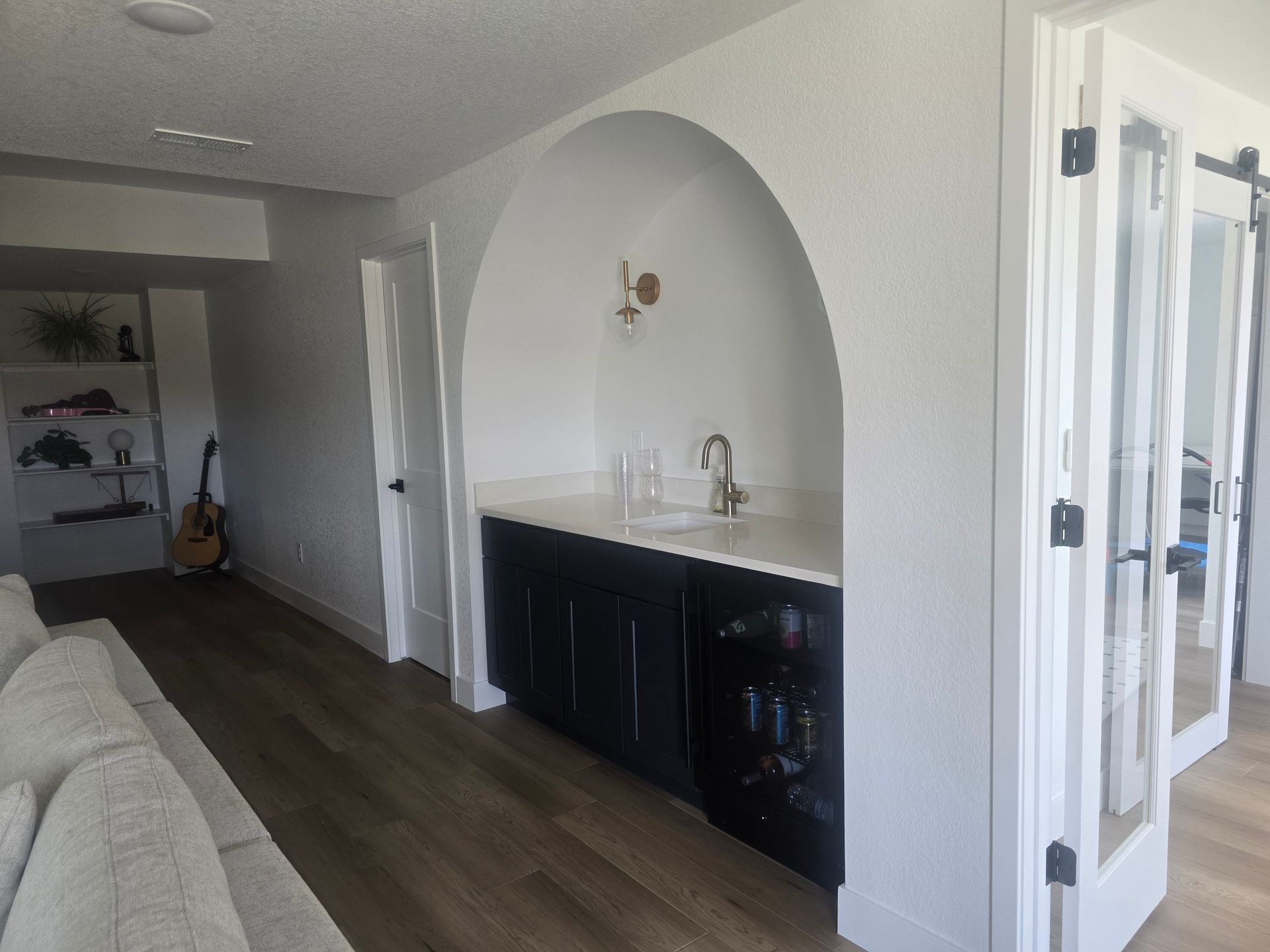 A home bar with a black cabinet, marble countertop, and arched alcove. Two French doors and a built-in shelf are to the right and left.
