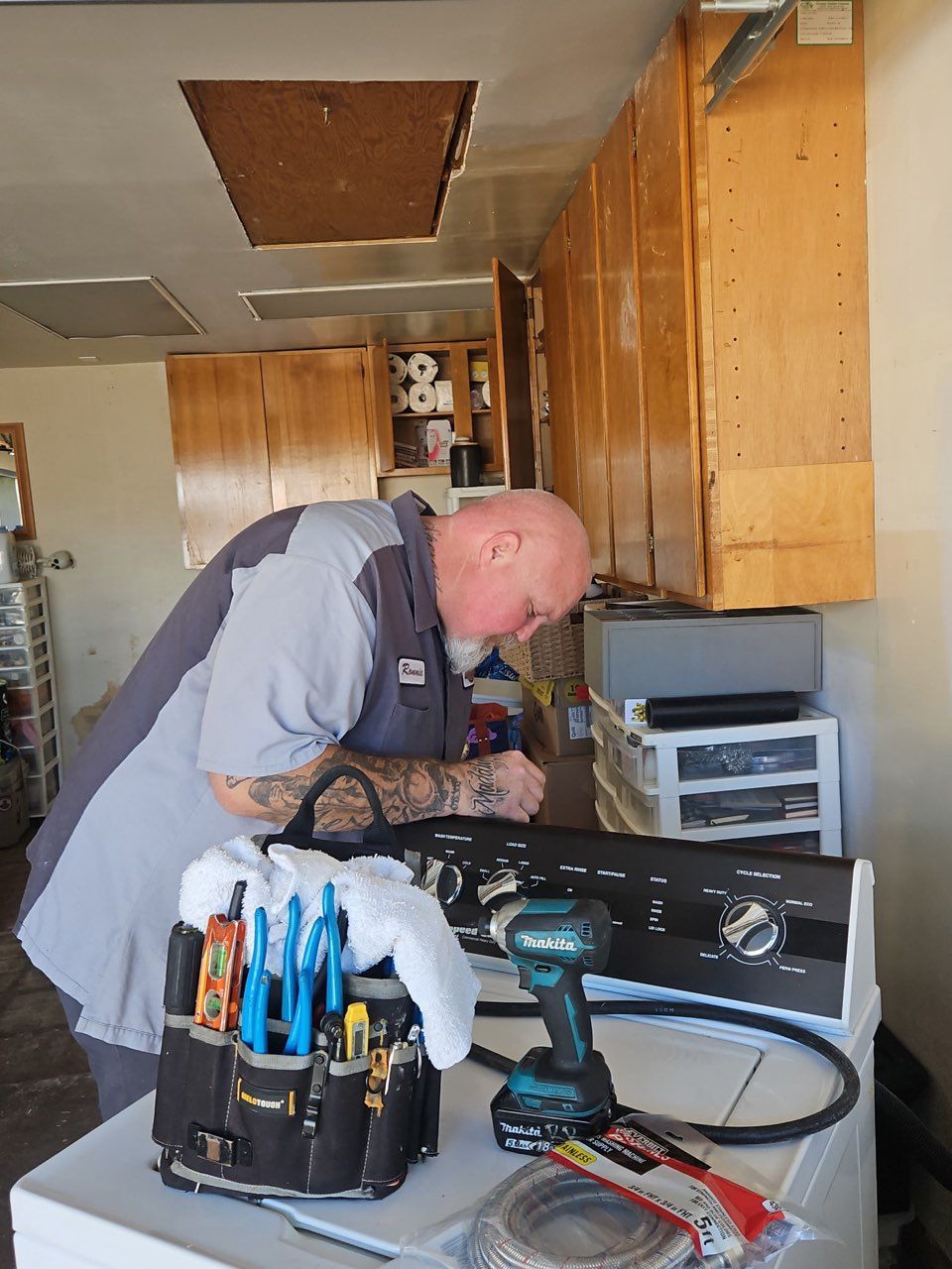 Technician leaning over an open appliance, using tools in a cluttered workshop kitchen.