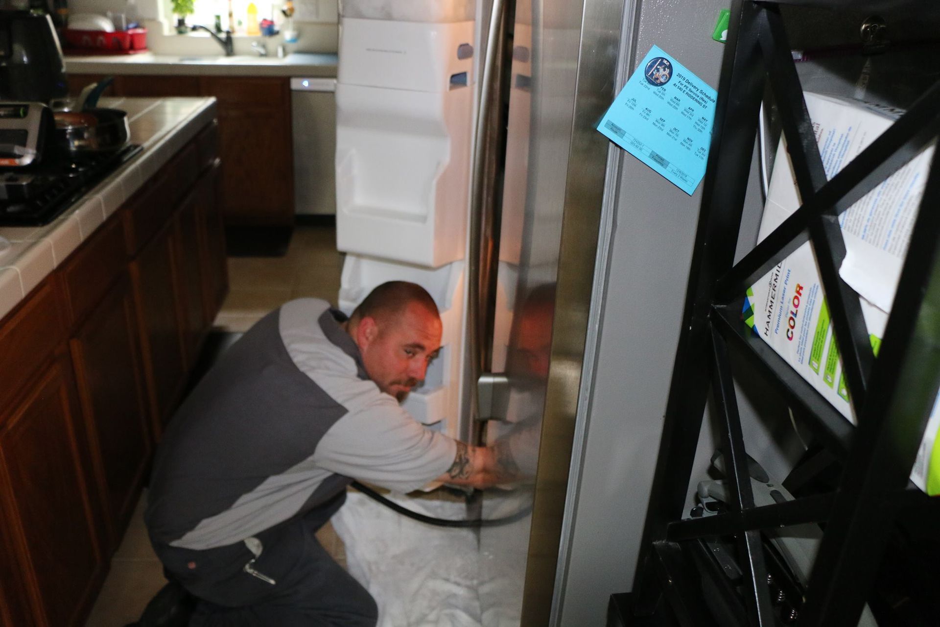 A technician in a grey shirt kneels in a kitchen to repair a stainless steel refrigerator.