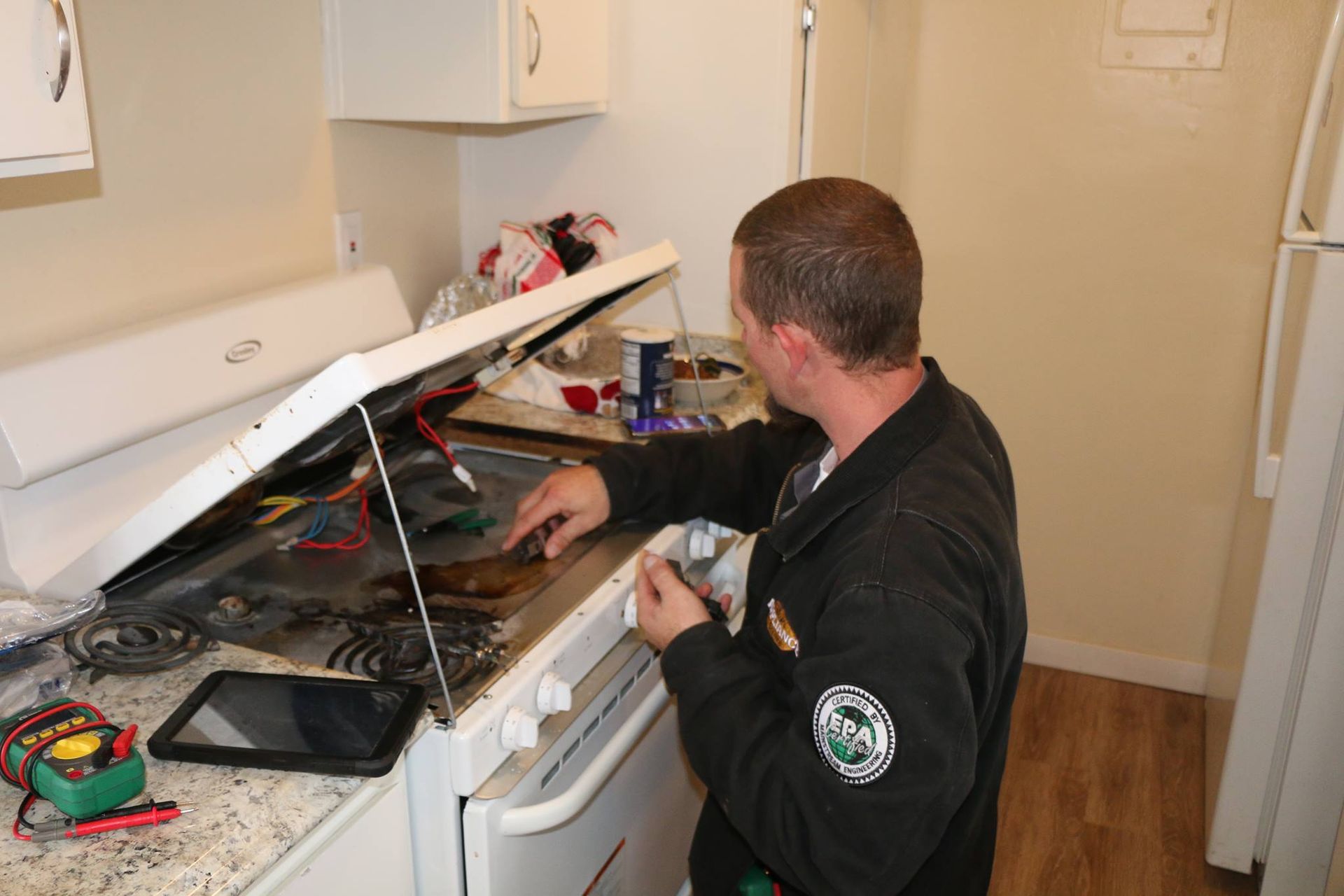 A technician works on the open top of an electric stove in a kitchen, using a multimeter nearby.