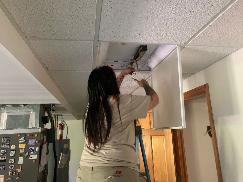 Person standing on a ladder fixing a ceiling light panel in a hallway.