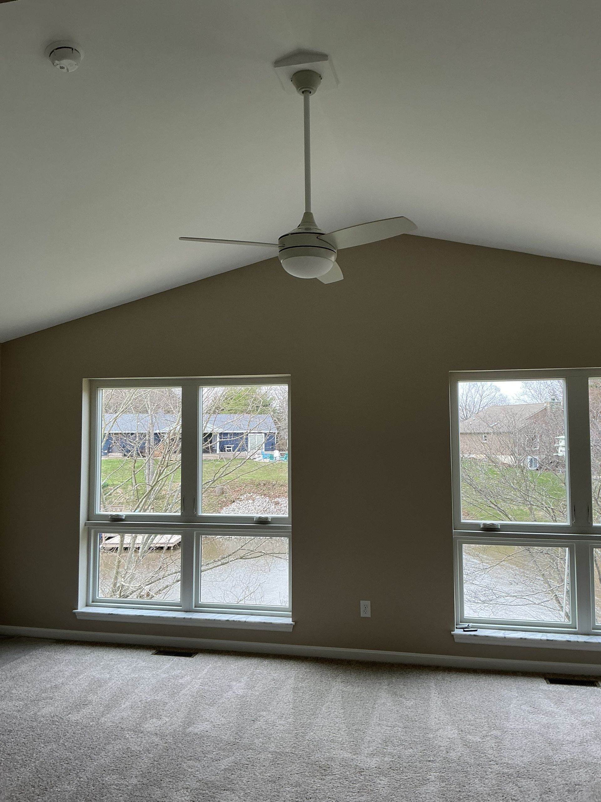 Empty beige room with two large windows, a ceiling fan, and carpeted floor.