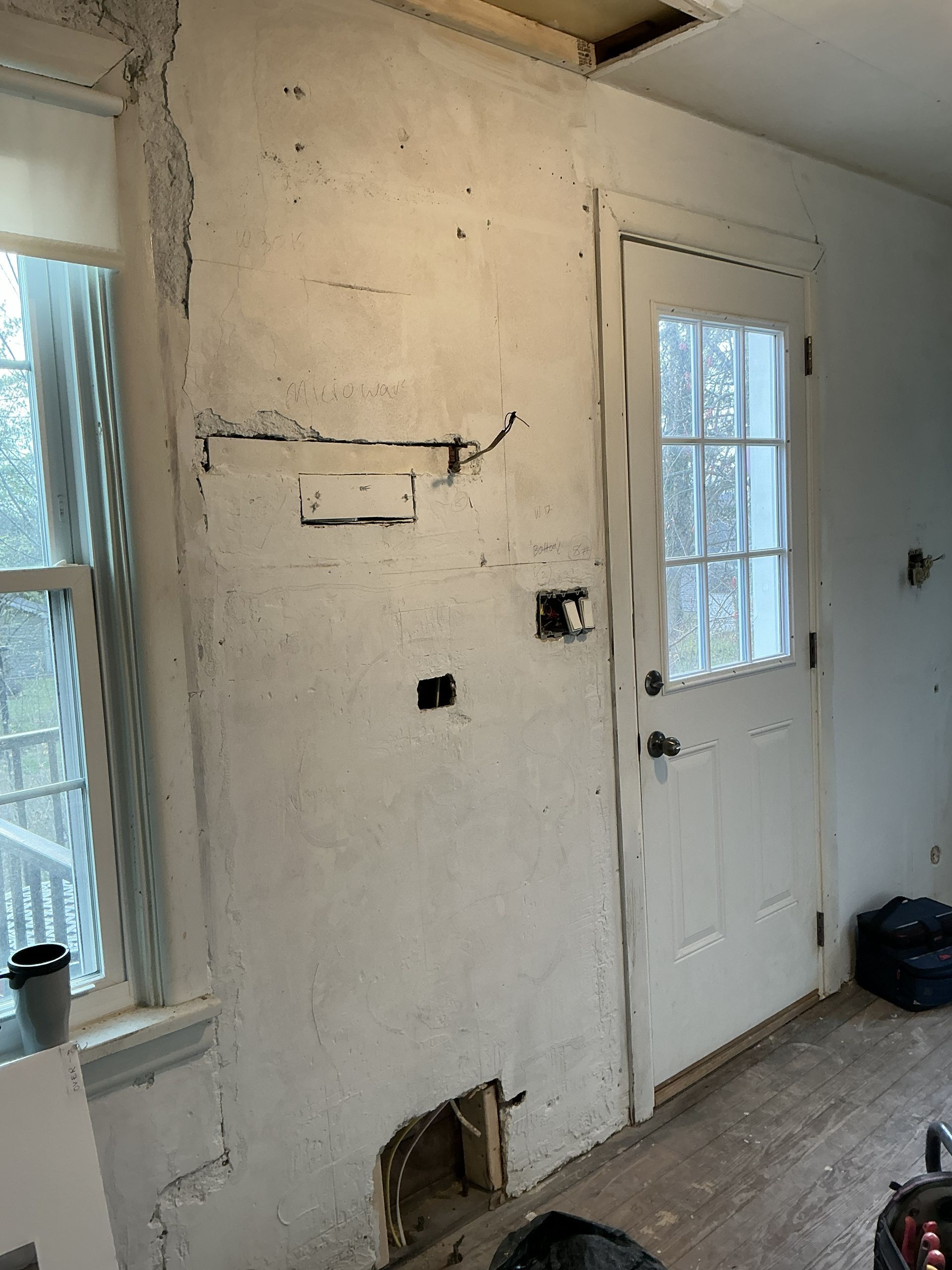 Damaged interior wall with exposed wiring beside a white door and windows in a small room