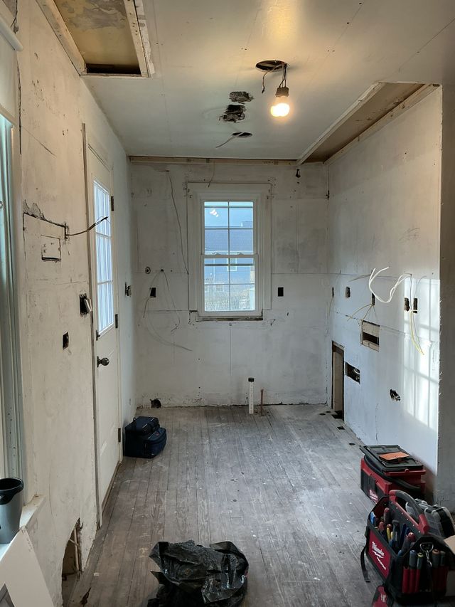 Narrow unfinished room with white walls, a window, exposed light fixture, and tools scattered on the floor.