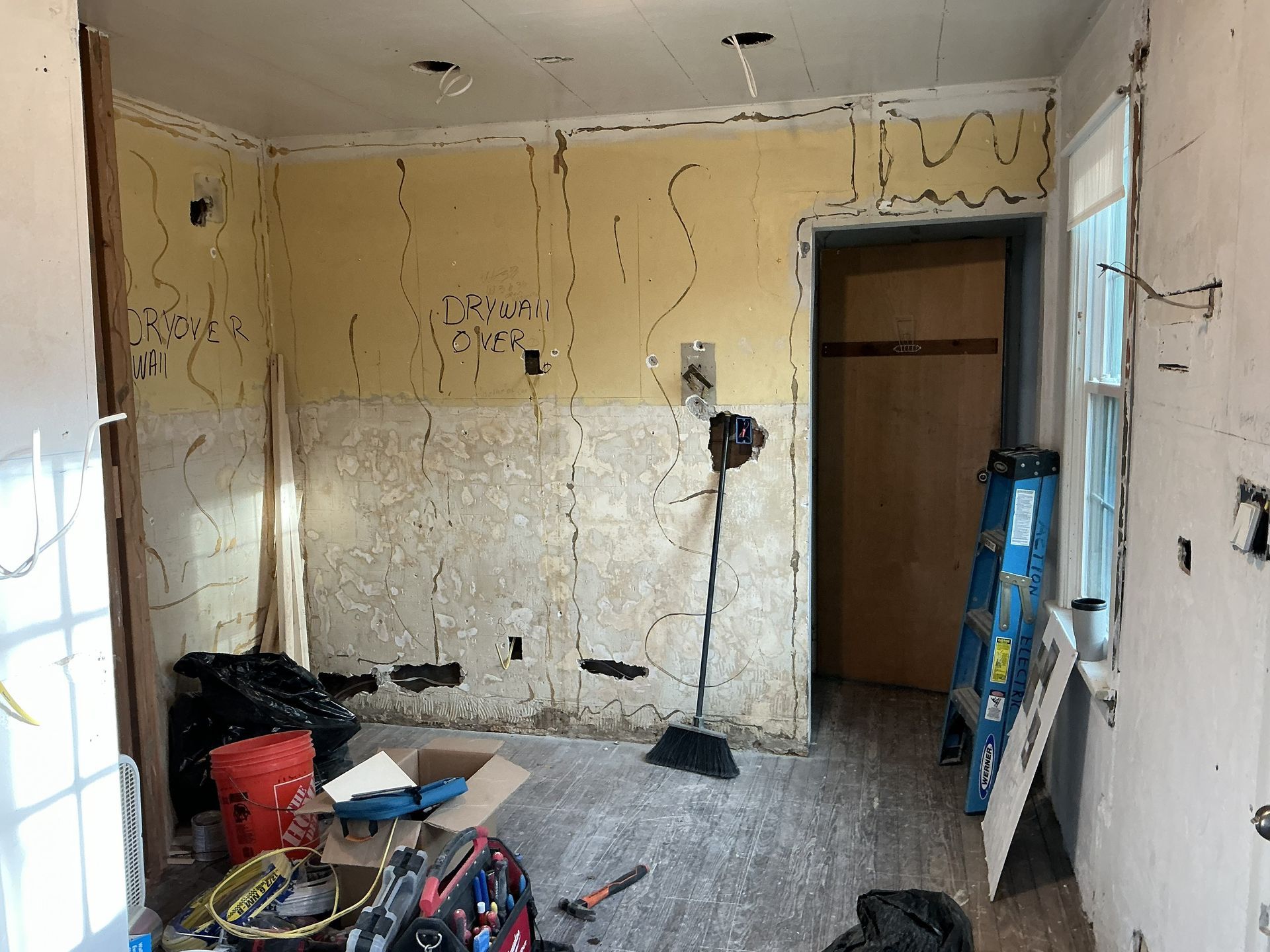 Bare room under renovation with peeling walls, debris on the floor, and a ladder by the doorway