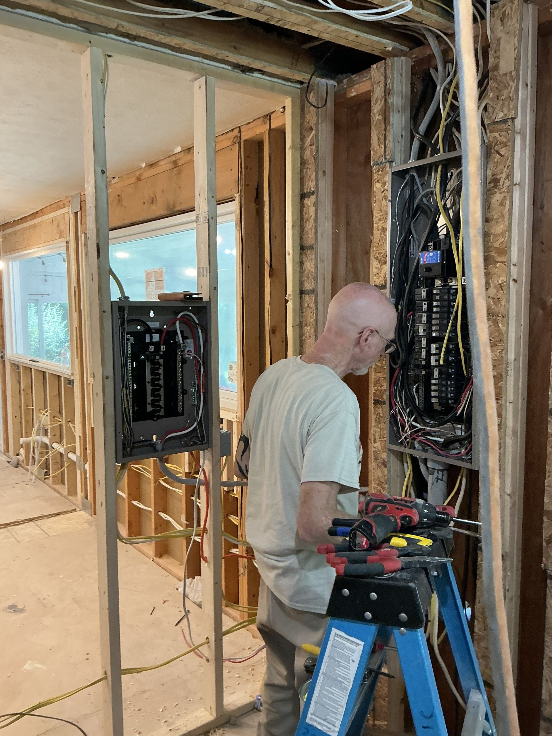 Worker wiring electrical panel in an unfinished room with exposed studs and tools on a step ladder