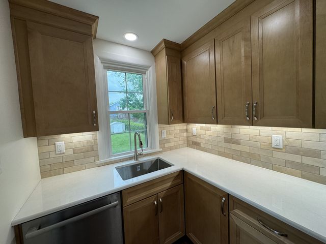 Modern kitchen corner with wood cabinets, white countertops, sink under a window, and tiled backsplash.
