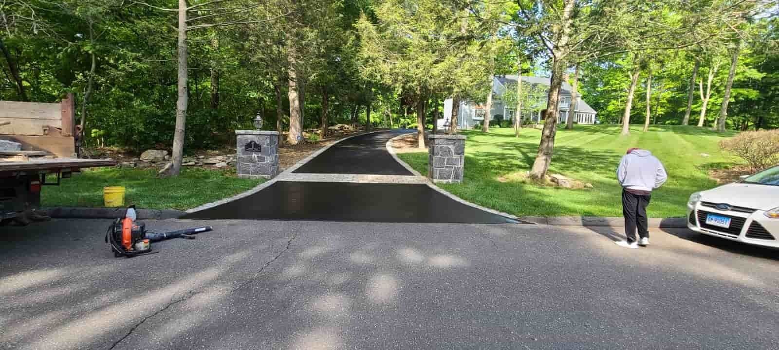 Driveway entrance with a black asphalt ramp, trees on both sides, and two people near the road.