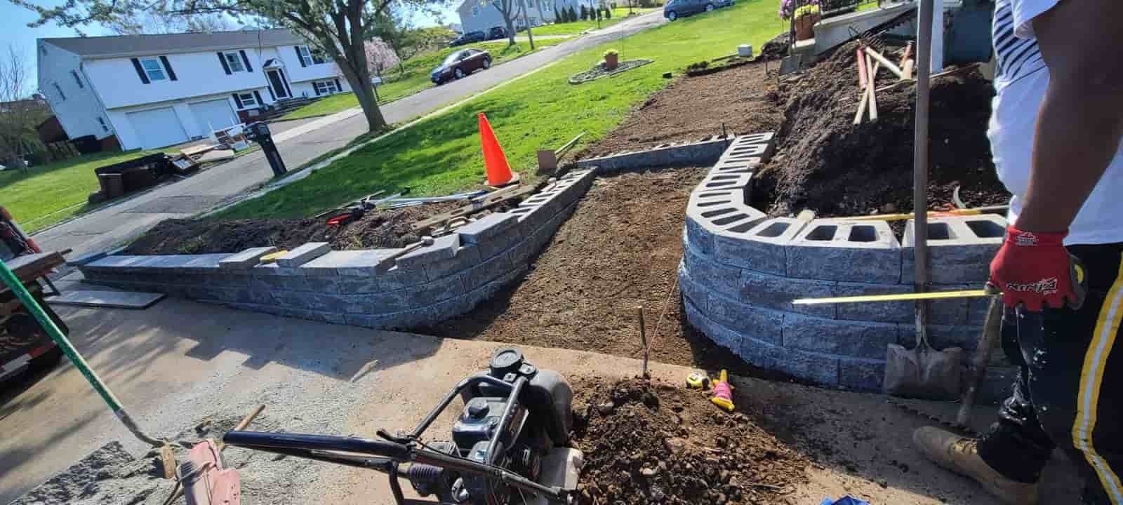 Workers build a curved stone retaining wall beside a sidewalk and a house, with tools and gravel visible.
