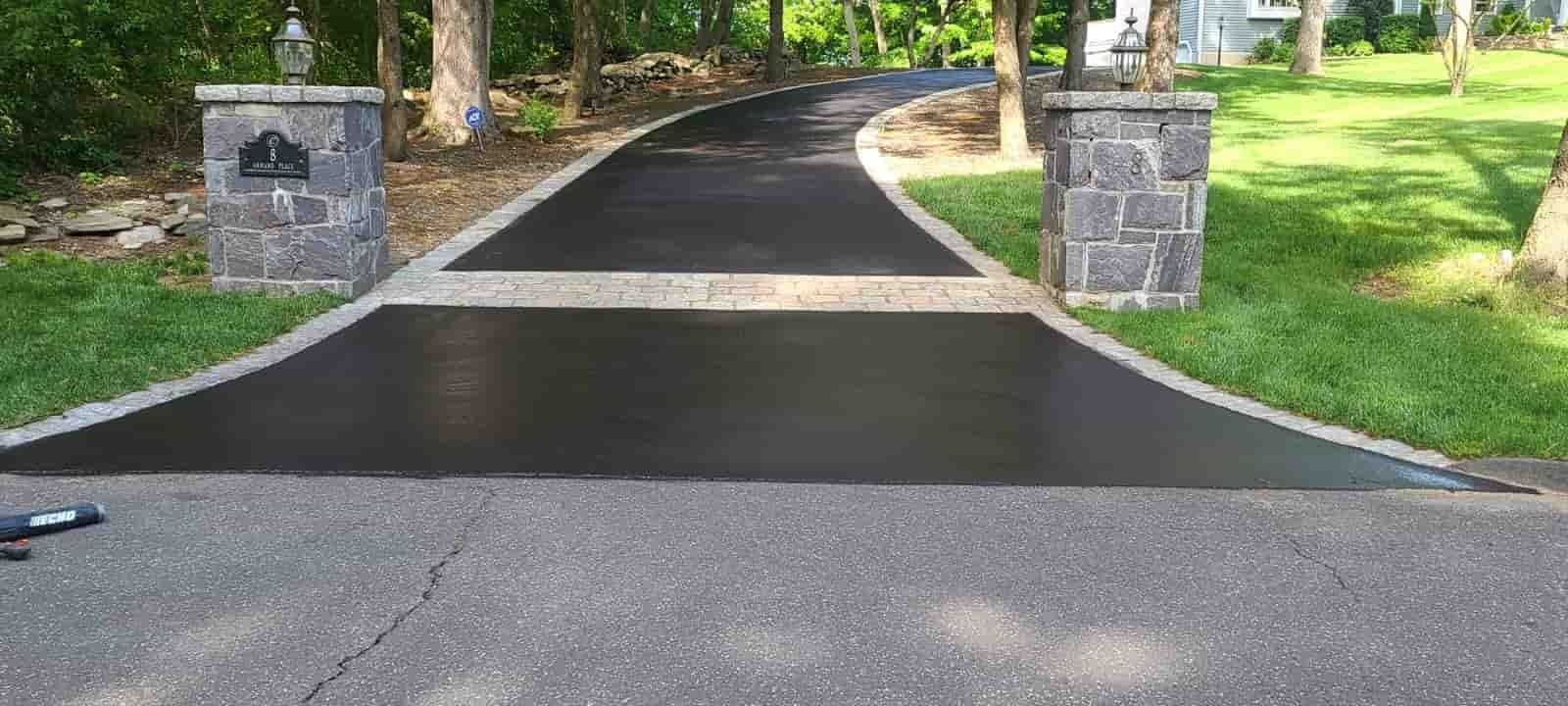 Paved driveway entrance between stone pillars, bordered by trees and green grass.