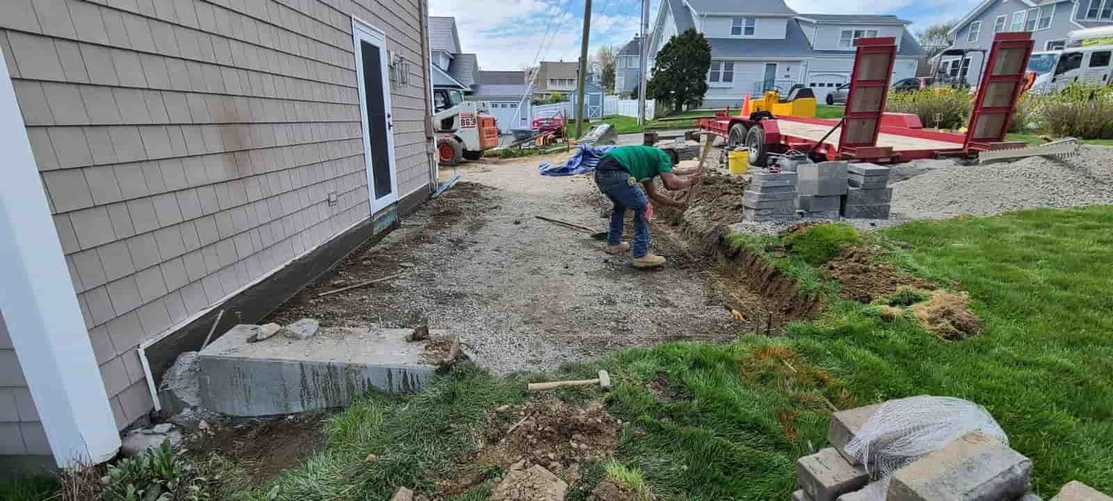 Worker shoveling gravel beside a house during outdoor construction, with a red excavator in the background