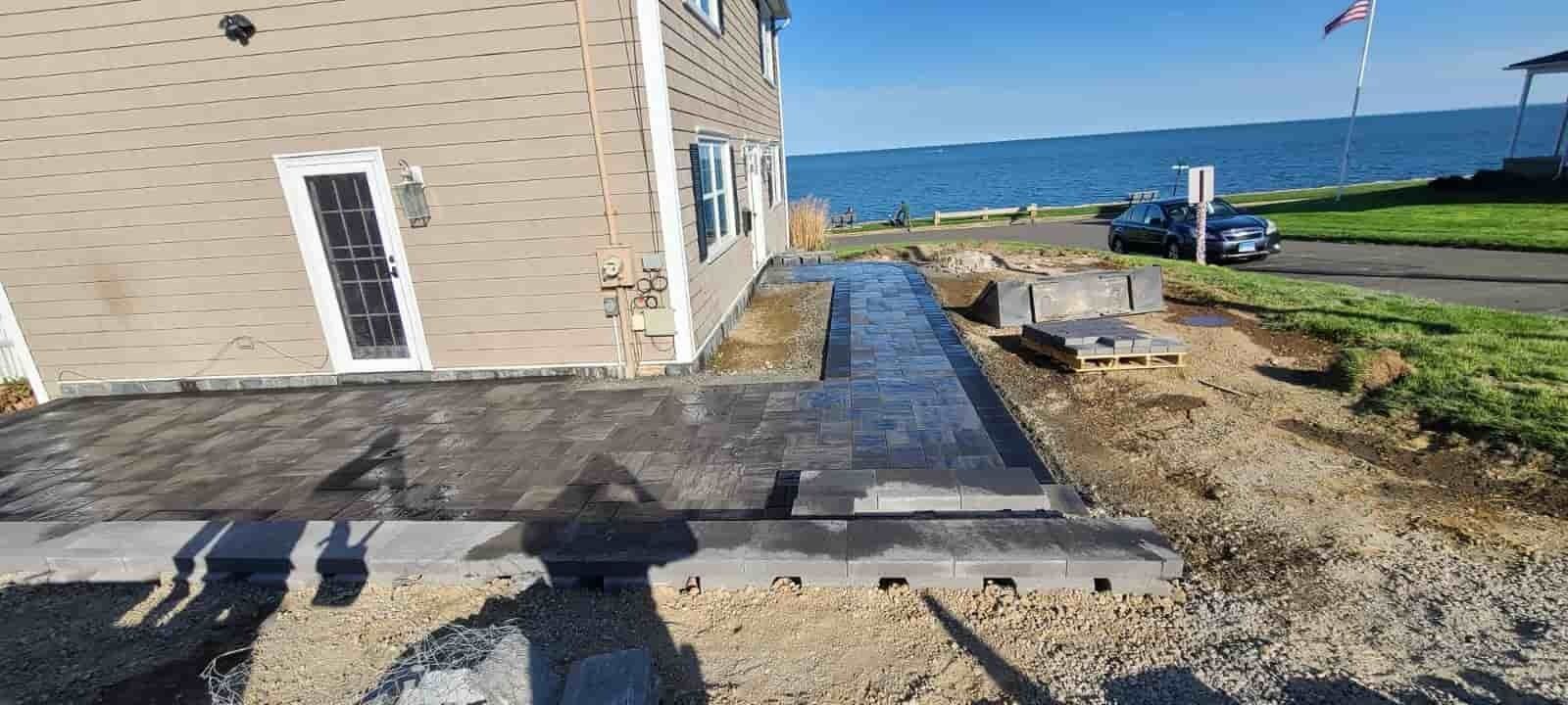 Coastal house side yard with a paved patio, gravel path, blue sea, and parked car under a sunny sky