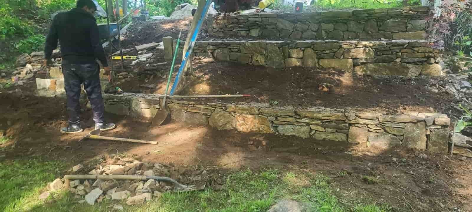 Person standing in a dirt yard beside a stone retaining wall and a blue post under trees