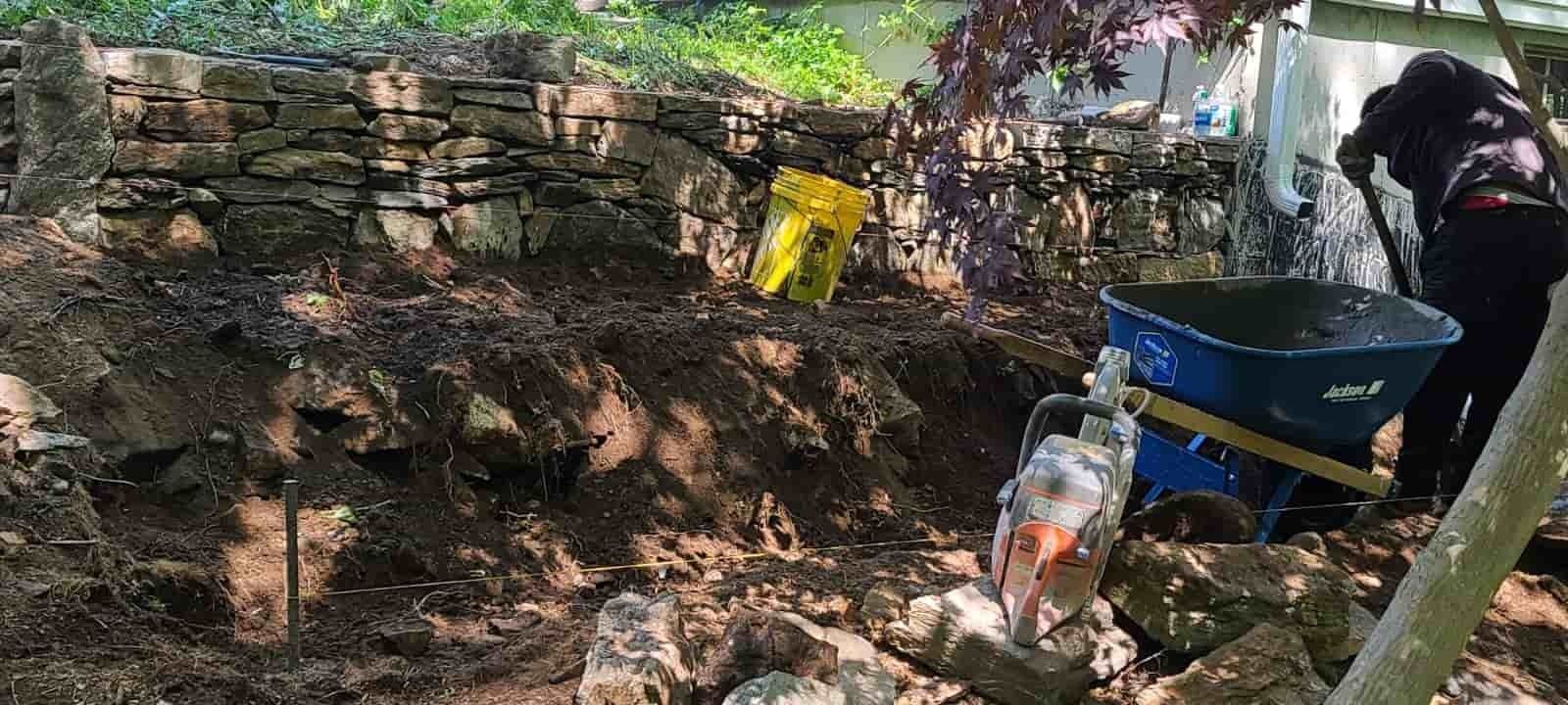 Person working in a rocky garden with a wheelbarrow and tools near a stone wall