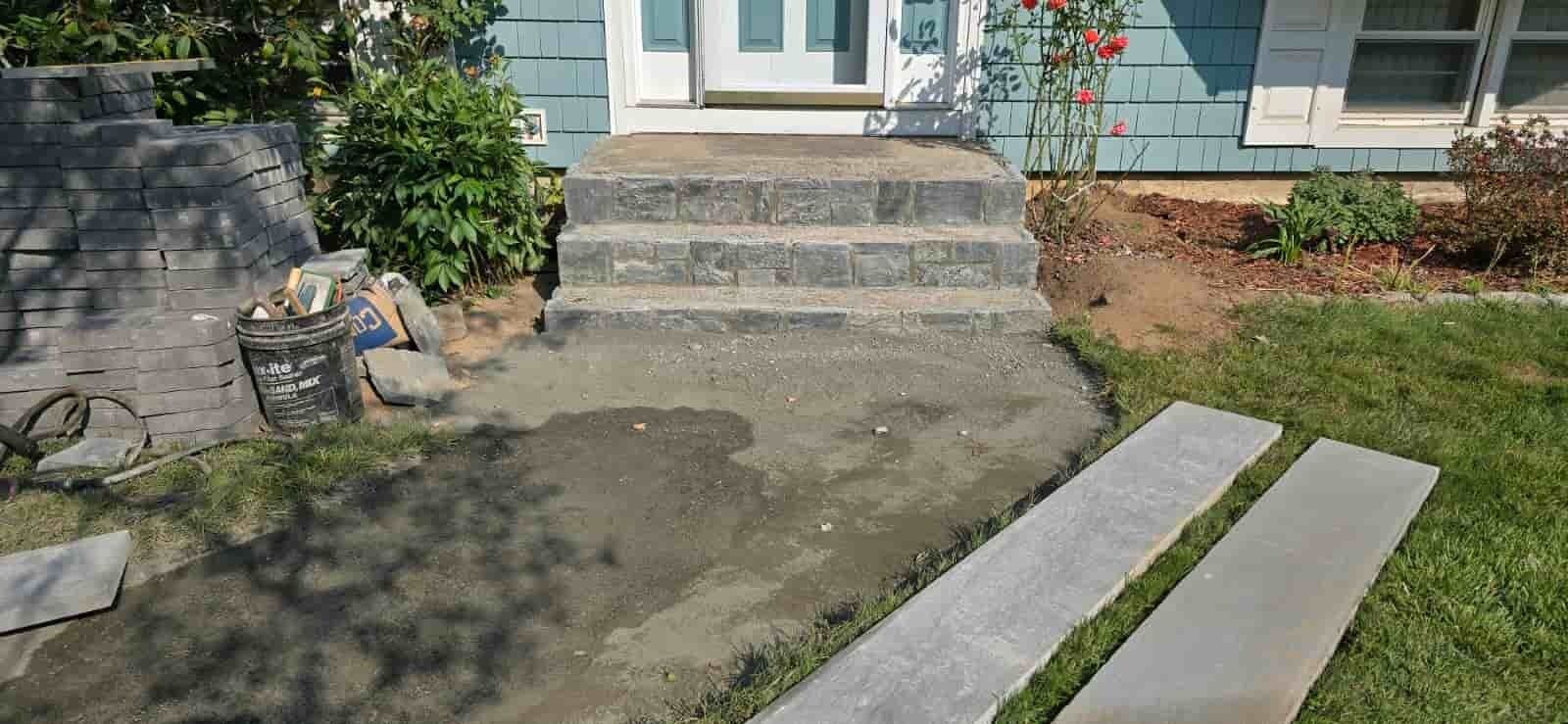 Front stoop with concrete steps, wet patio, and two narrow garden pavers leading to a house entrance.
