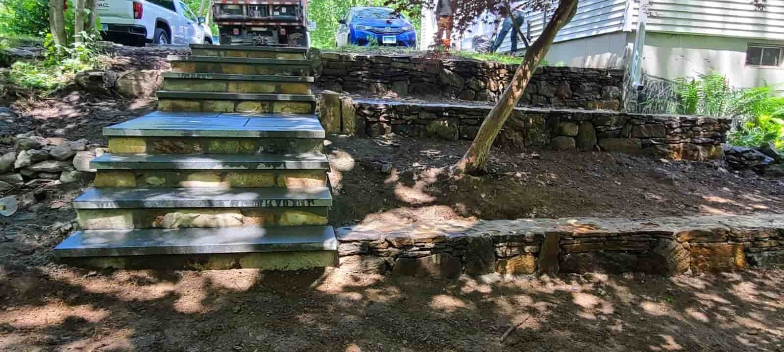 Stone steps in a shaded park leading up to a grassy area with trees and parked cars