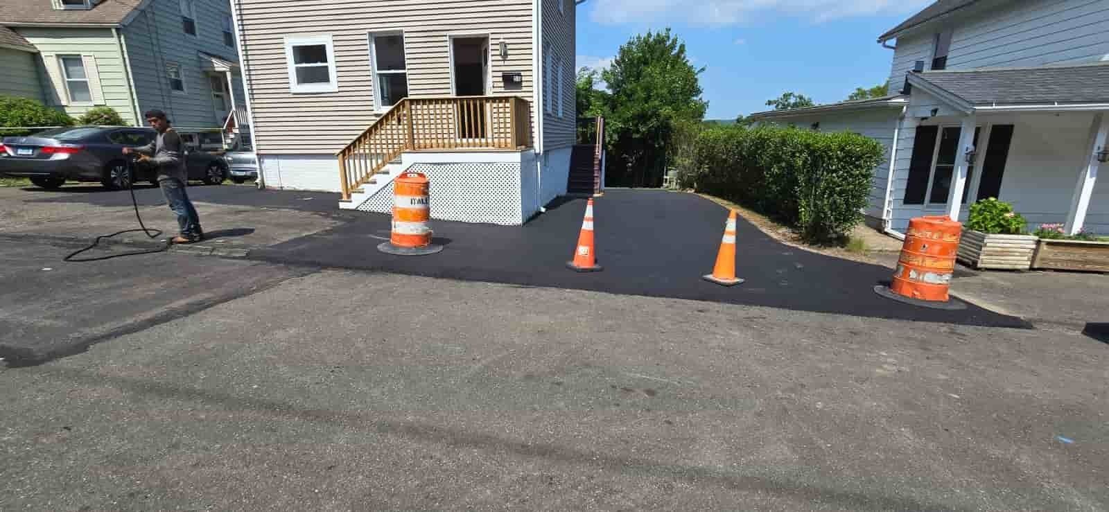 Orange traffic cones block a driveway beside a white house and street, with a dark car on the left.