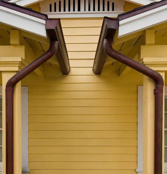 A symmetrical close-up of a yellow house exterior showing two brown gutters mounted to white pillars under sloped roofs.