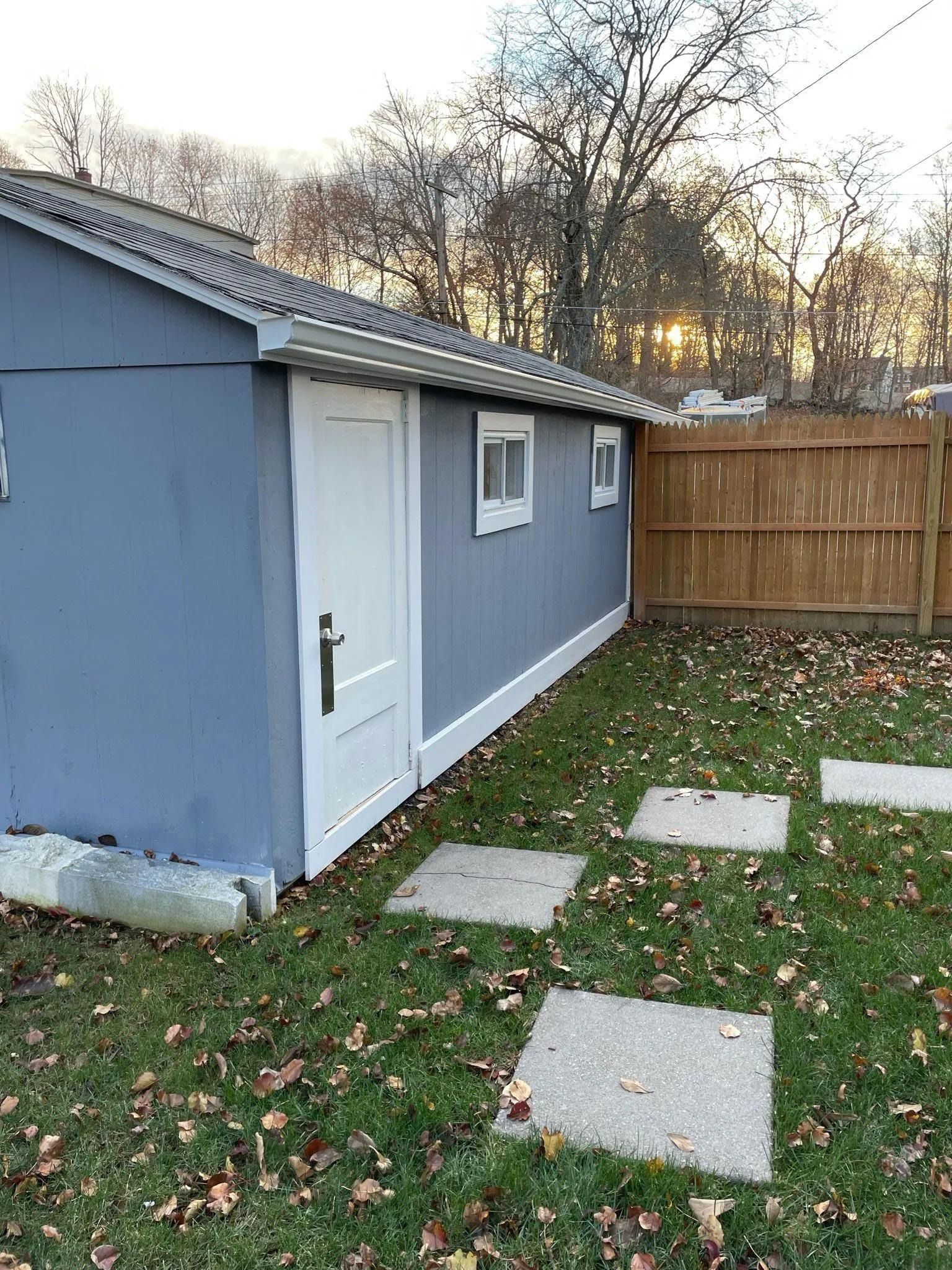 A grey backyard shed with a white door and windows, situated next to a wooden fence on a grassy lot with stone pavers.