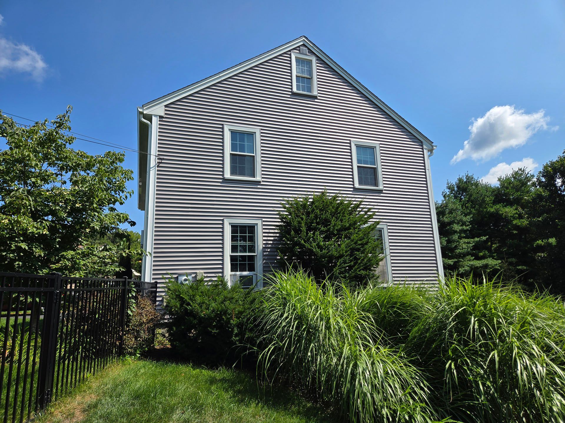 Gray clapboard house with white trim, black fence, and tall grass under a blue sky.