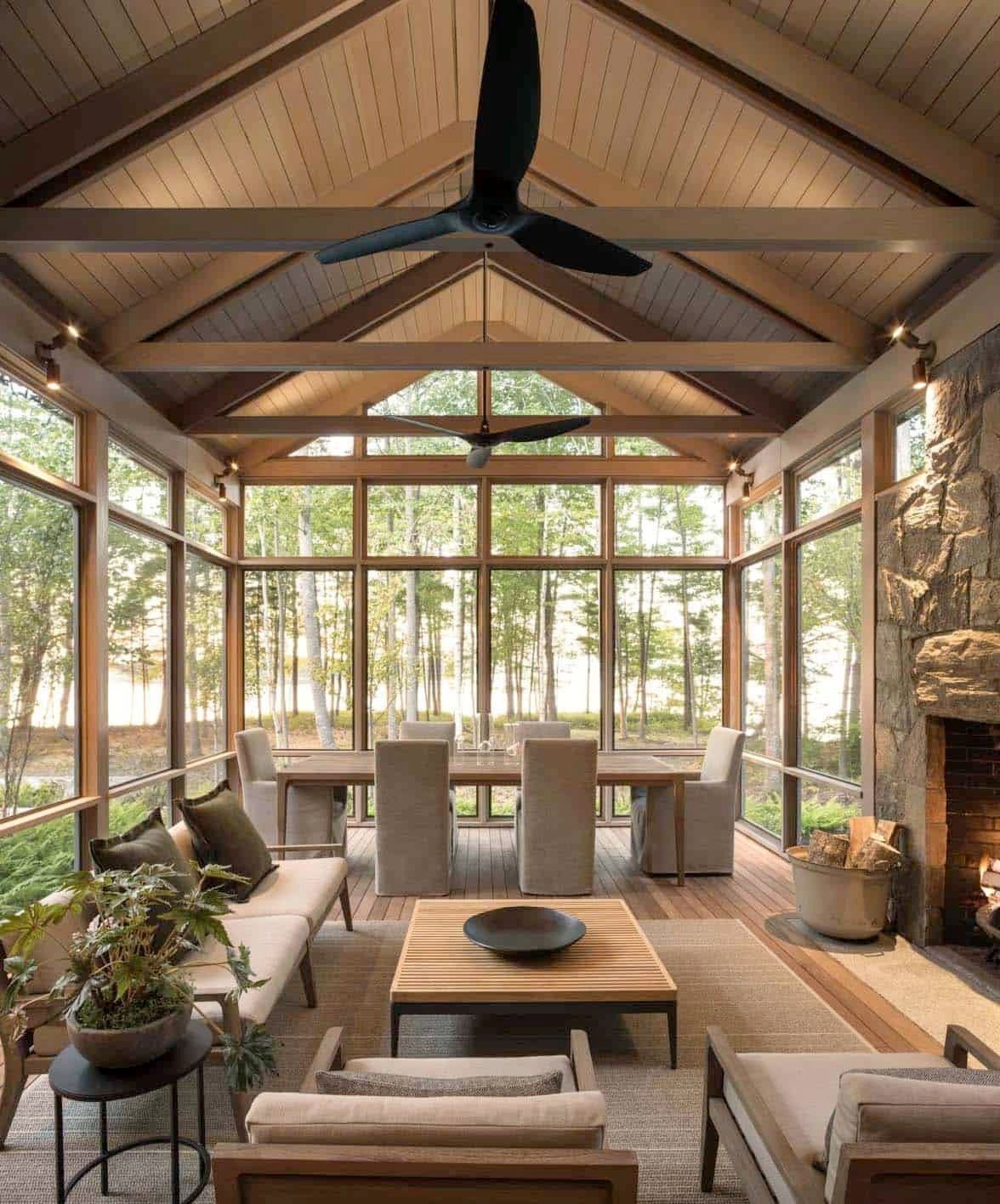 Screened-in porch with wood beams, stone fireplace, and modern furniture overlooking trees and lake.