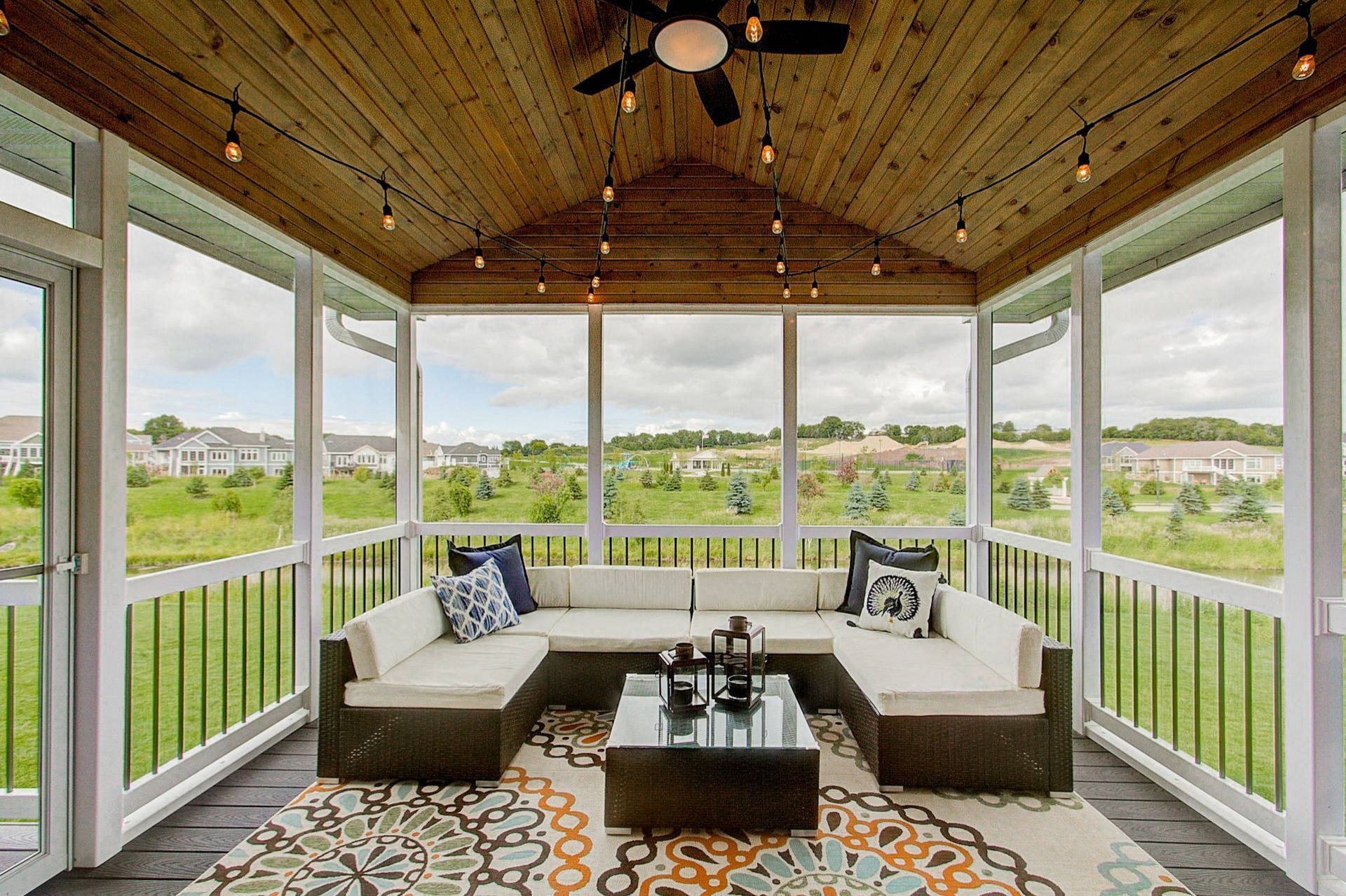 Screened-in porch with U-shaped sofa, rug, and coffee table. Wooden ceiling with string lights, overlooking a grassy landscape.