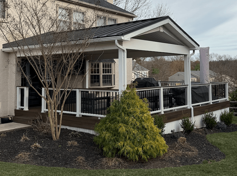 Covered deck with black railing and metal roof attached to a beige house, surrounded by landscaping.