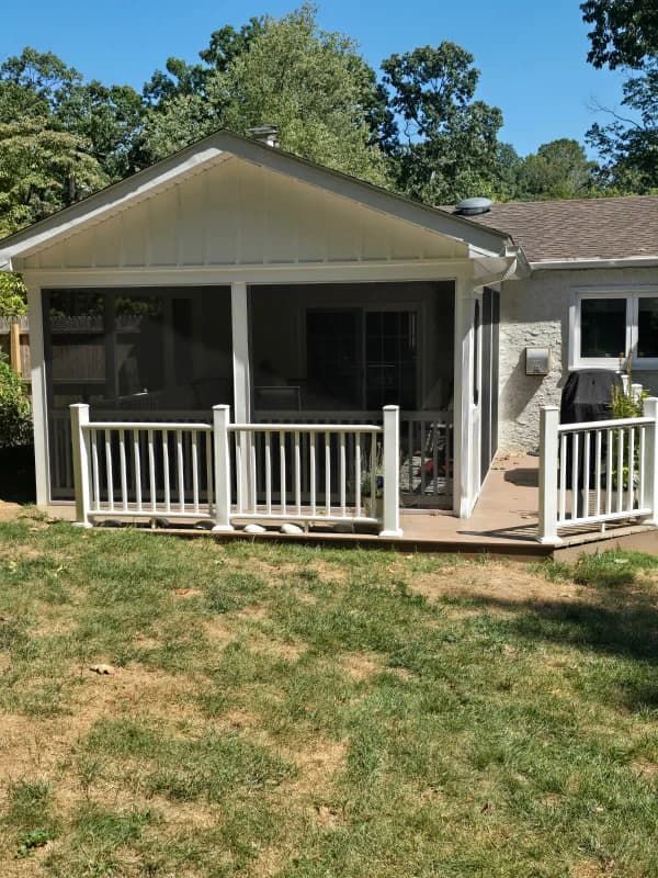 Screened porch with white railings and roof, attached to a house with a concrete wall, on a grassy lawn.