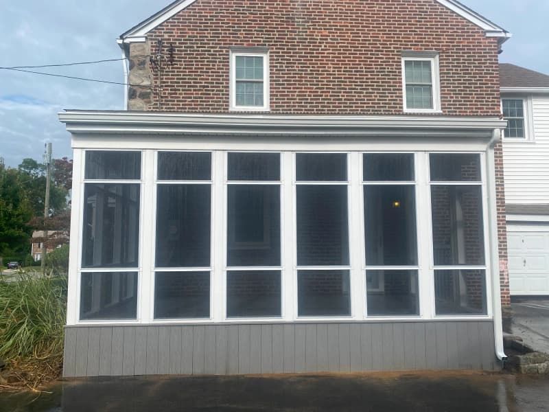 Screened-in porch addition with gray siding, white frames, and a brick house in the background.