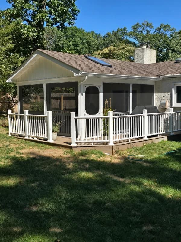 Screened-in porch attached to a house with white railing. Green grass and trees surround it.
