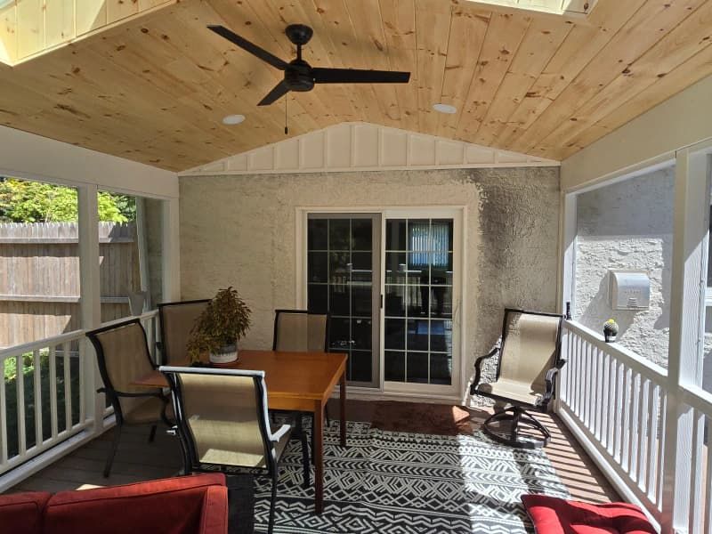 Screened porch with dining table, chairs, and ceiling fan; wooden ceiling, stucco walls, and outdoor rug.