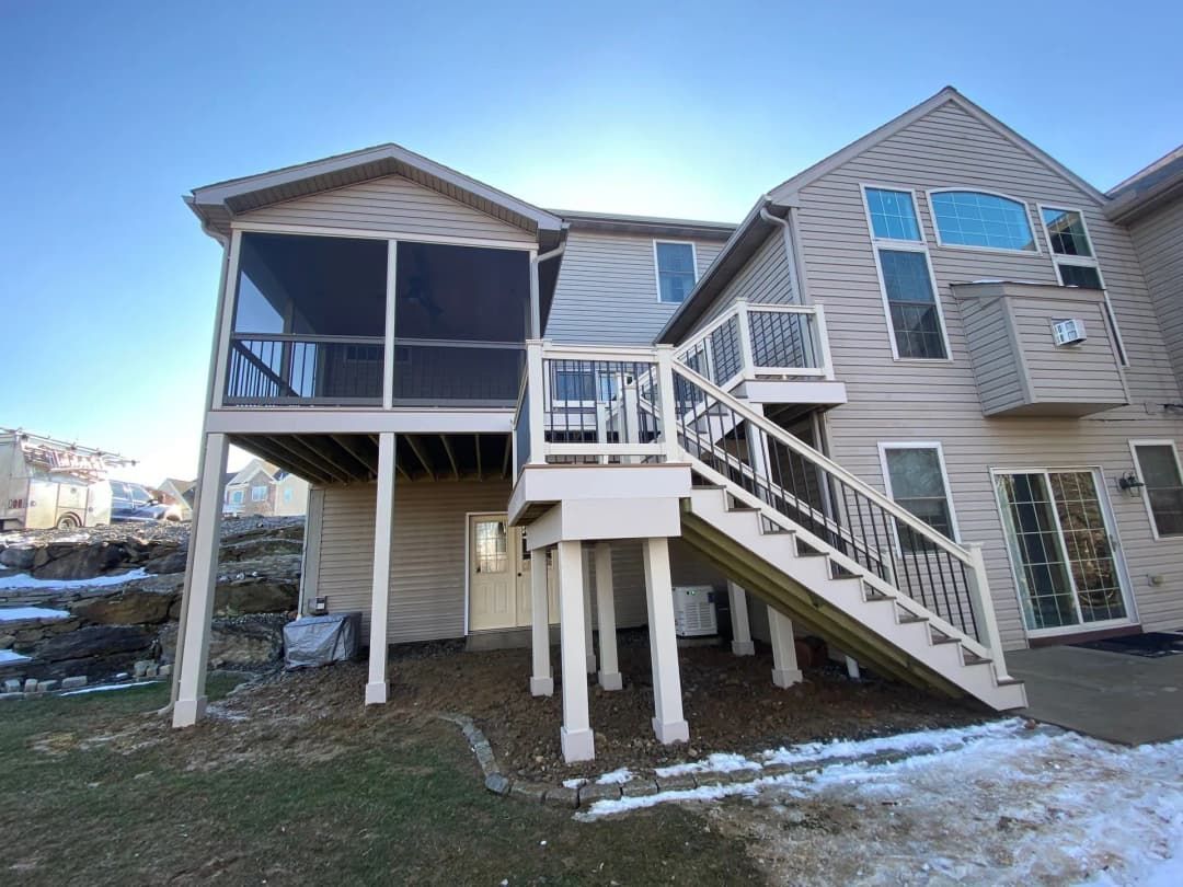 Beige two-story house with elevated wooden deck and stairs. A screened porch sits on the deck. Winter setting.