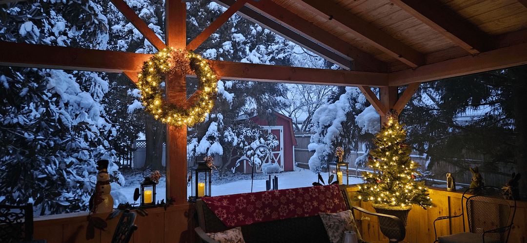 A cozy, snow-covered porch decorated with Christmas lights and a wreath, overlooking a snowy yard.