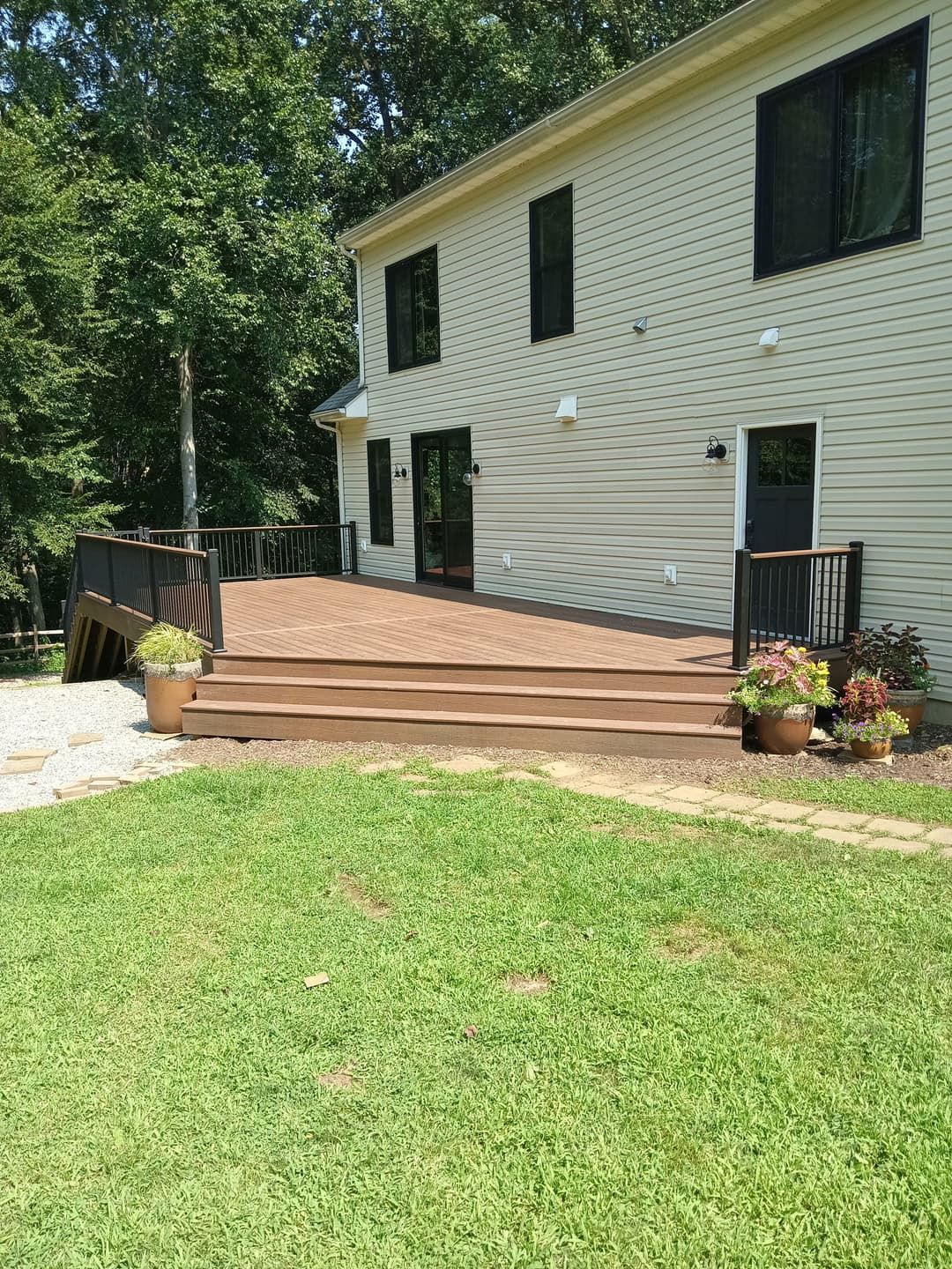 Back of a two-story beige house with a brown deck. Green grass in the foreground, trees in the background.