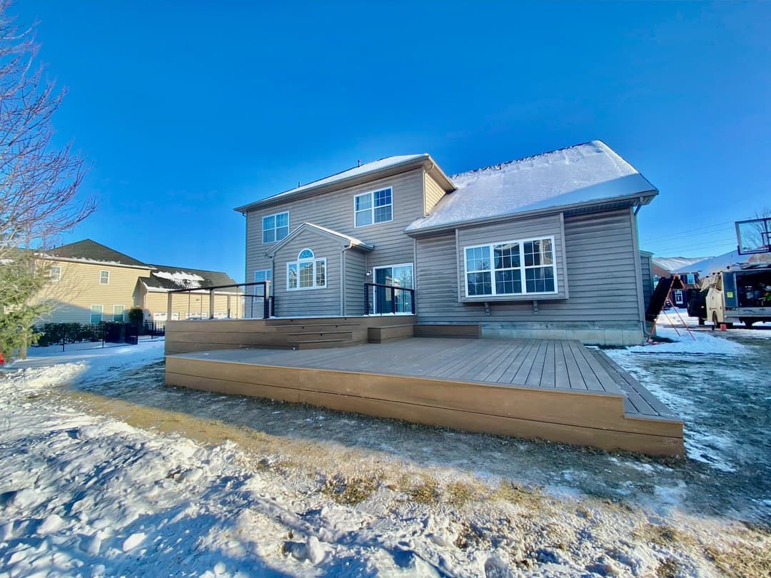 Back of a two-story house with a wooden deck, covered in snow on a sunny day.